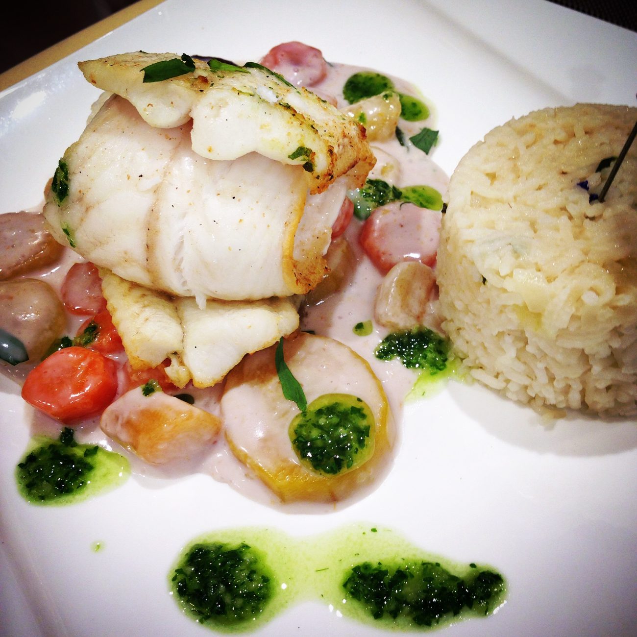 Un filet de poisson doré sur un lit de légumes, riz en timbale, sauce crémeuse et touches de pesto vert, dressé sur une assiette carrée blanche
